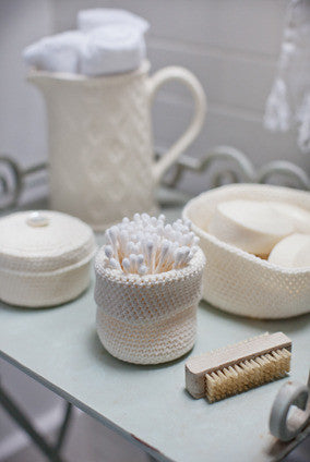 A set of crocheted baskets in various sizes made with a beige cotton yarn, displayed on a table with a vase, towels, and a brush.